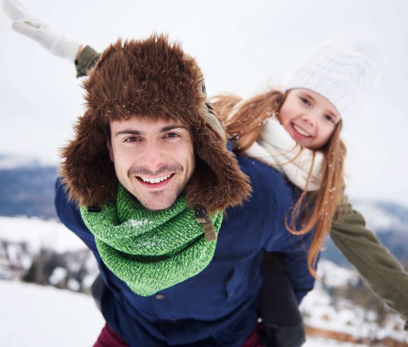 Lächelnder Vater mit Fellmütze trägt seine Tochter auf dem Rücken im verschneiten Winterwunderland. Perfekt für den Ratgeber 